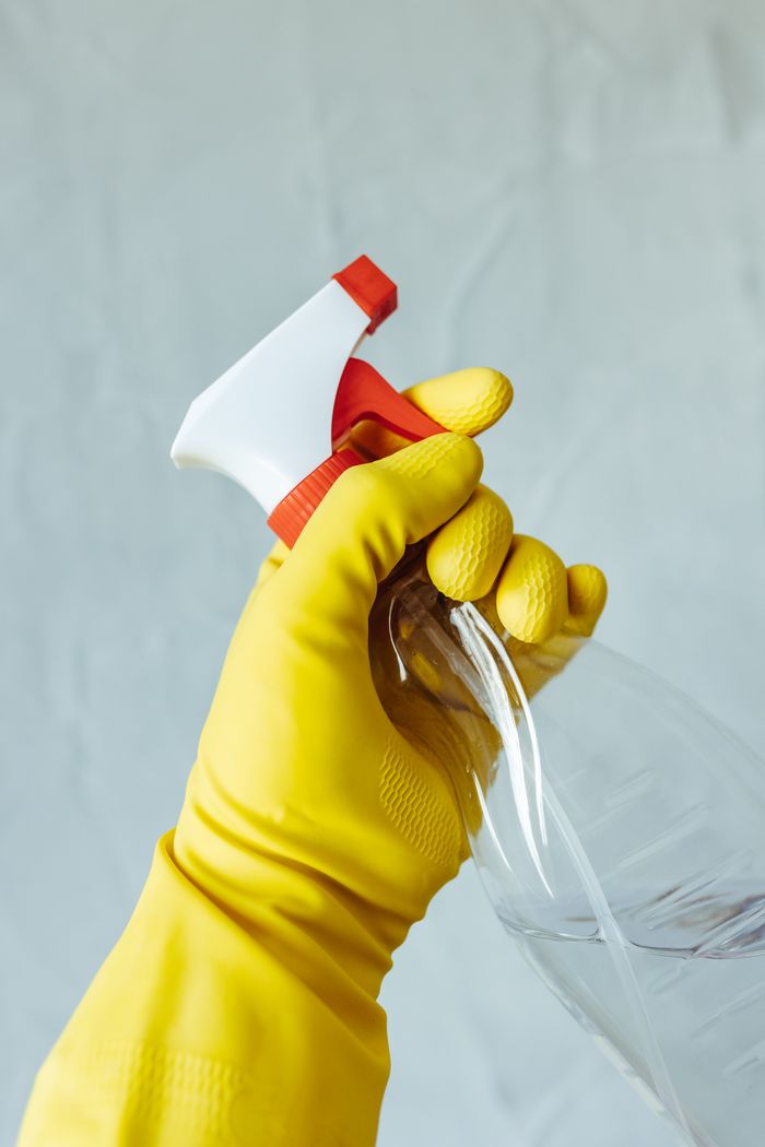 project-02-d A hand wearing a yellow rubber glove holds a red and white spray bottle against a neutral background.